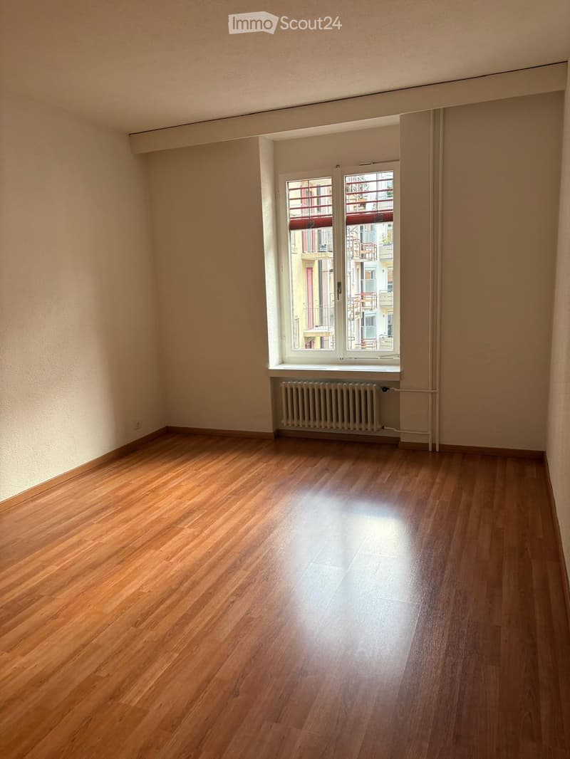 Empty room with wooden flooring, light-colored walls, and a window with a radiator below it.