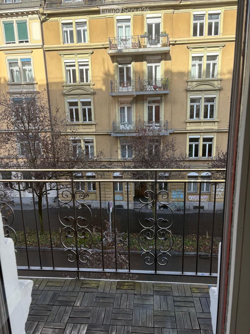 Balcony with wooden decking, ornate metal railing, overlooking a multi-story building with balconies and windows.