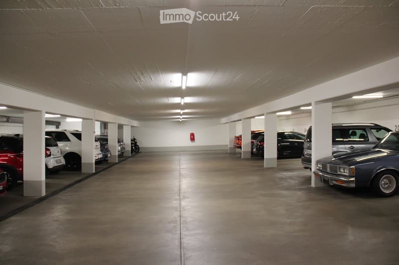 Underground parking garage with multiple parked cars, concrete floors, white pillars, and fluorescent lighting.