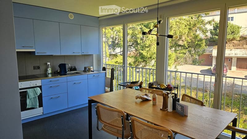 Modern kitchen with light blue cabinets, stainless steel appliances, and a wooden dining table with chairs, overlooking a balcony with outdoor seating.