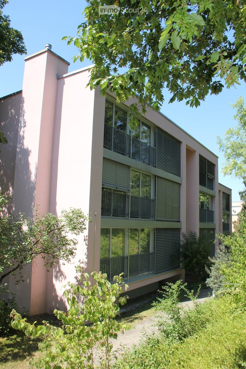 Modern two-story building with pink walls, large windows, and balconies surrounded by greenery and trees.