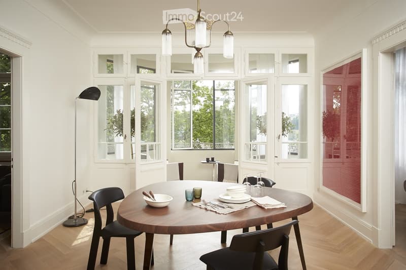 Spacious dining room with large windows overlooking a balcony or terrace, wooden oval dining table, black chairs, and a decorative chandelier above the table.