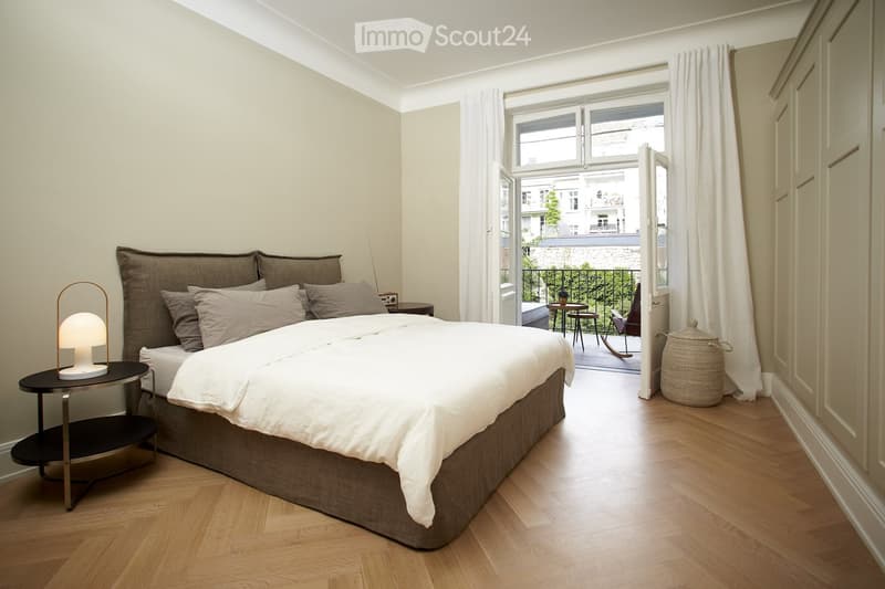 Spacious bedroom with large windows overlooking a balcony, hardwood floors, a king-size bed with a brown upholstered frame, and a side table with a lamp.