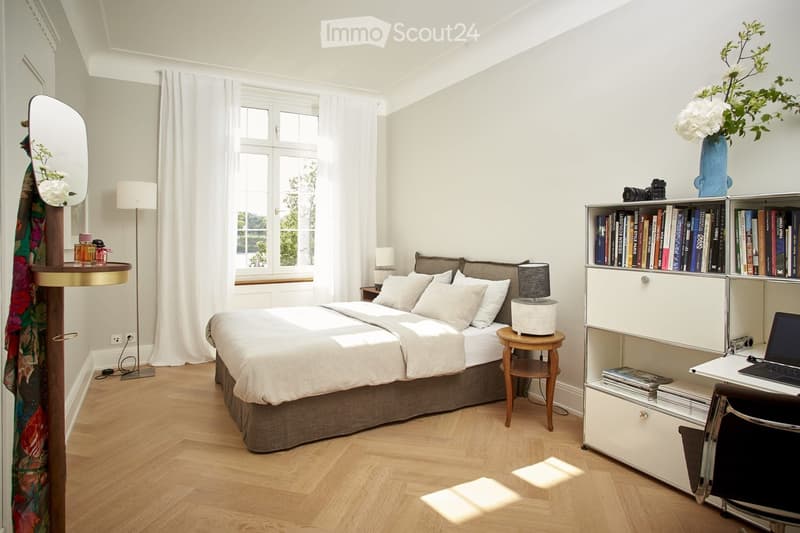 Spacious bedroom with large windows, hardwood flooring, a queen-size bed with neutral bedding, a bookshelf, and a desk with a laptop. The room has a cozy and bright atmosphere.