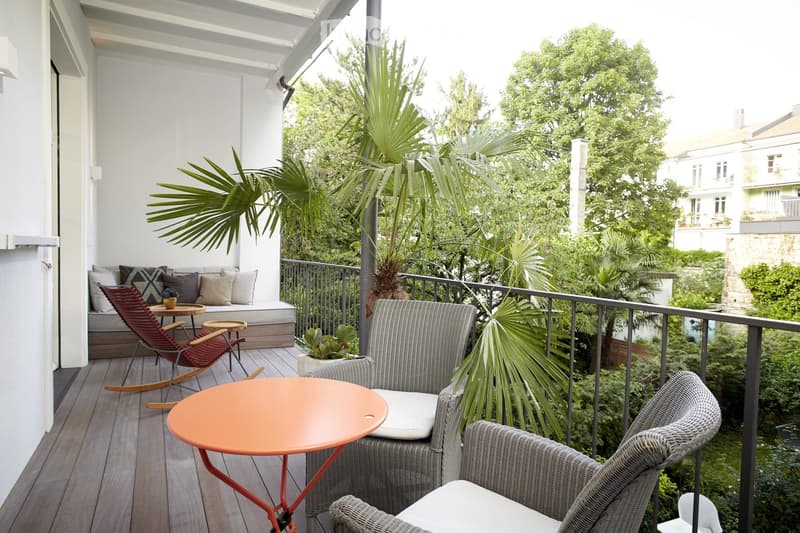 Covered balcony with wicker furniture, orange table, and lush tropical plants overlooking a green garden area