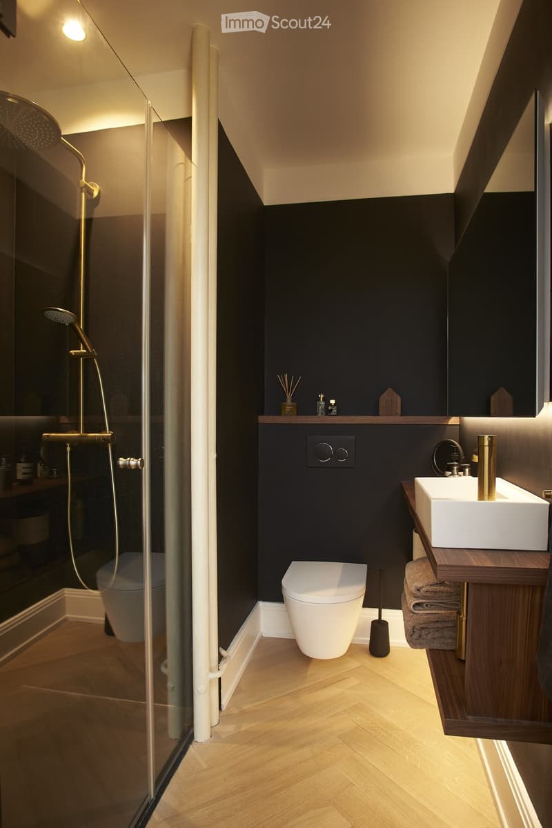 Modern bathroom with black walls, white toilet, vessel sink on wooden vanity, and glass shower enclosure with gold fixtures