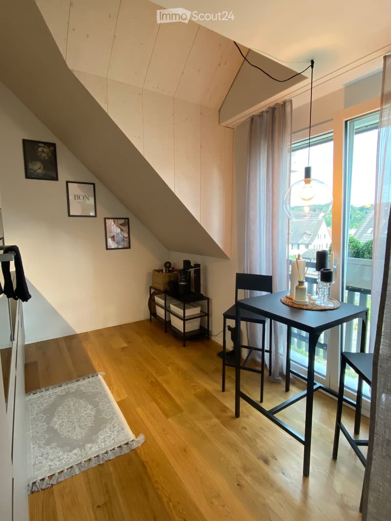 A cozy attic room with wooden flooring, large windows, and modern decor including a table, chairs, and a hanging lamp.
