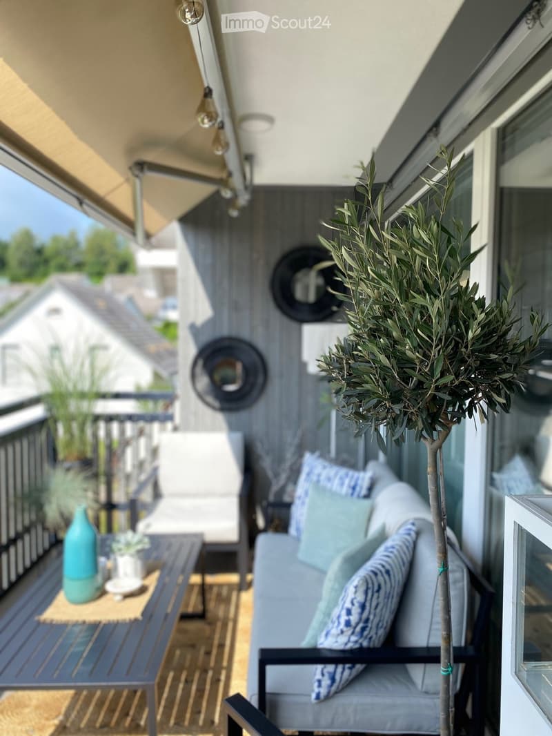Balcony with a grey sofa, blue and white patterned cushions, a small table with a blue vase, and a potted olive tree.