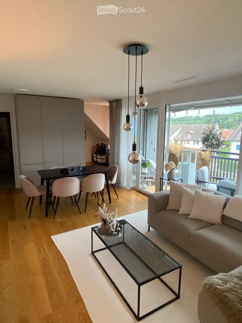 Modern living and dining area with light wooden flooring, a grey sofa, glass coffee table, dining table with chairs, pendant lights, and large windows with a view of the garden.