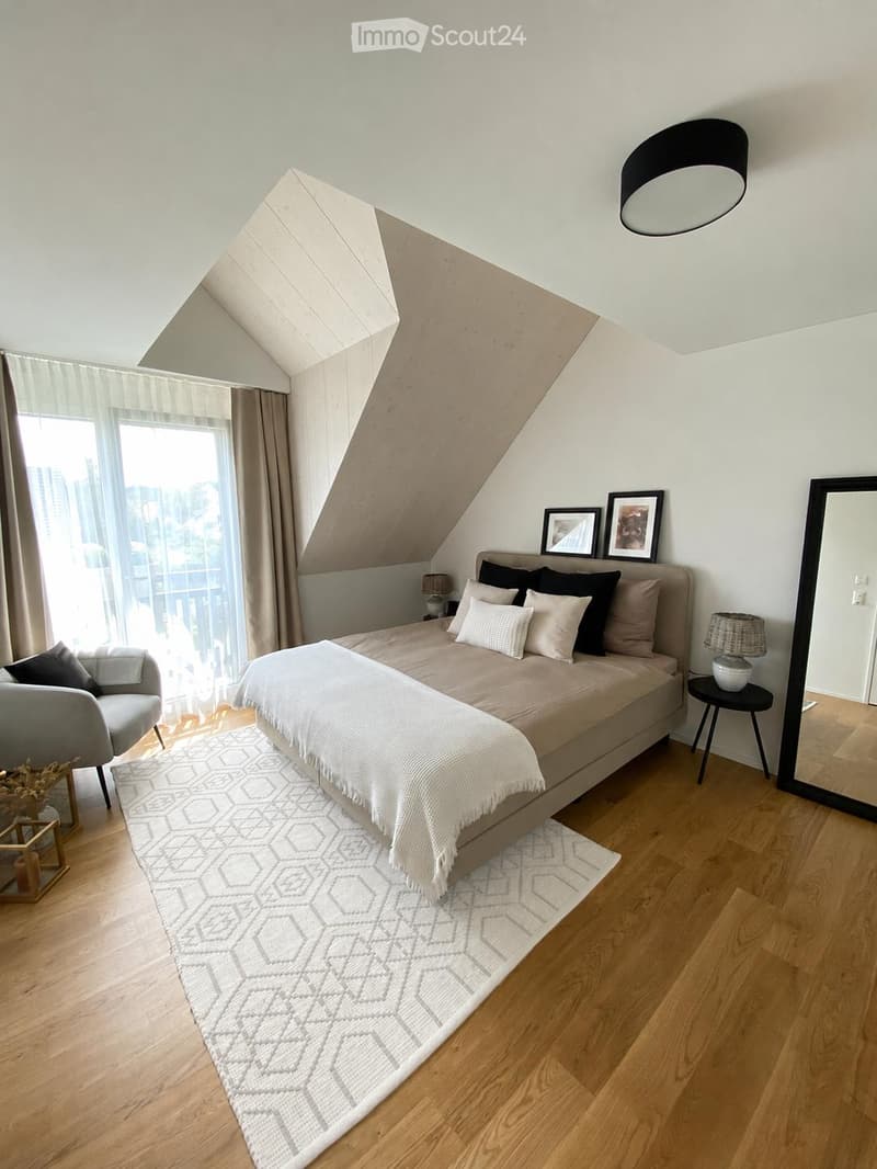 Modern bedroom with a large bed, grey armchair, side table, mirror, and decorative picture frames. The room features wooden flooring, a white rug, and large windows with city views.