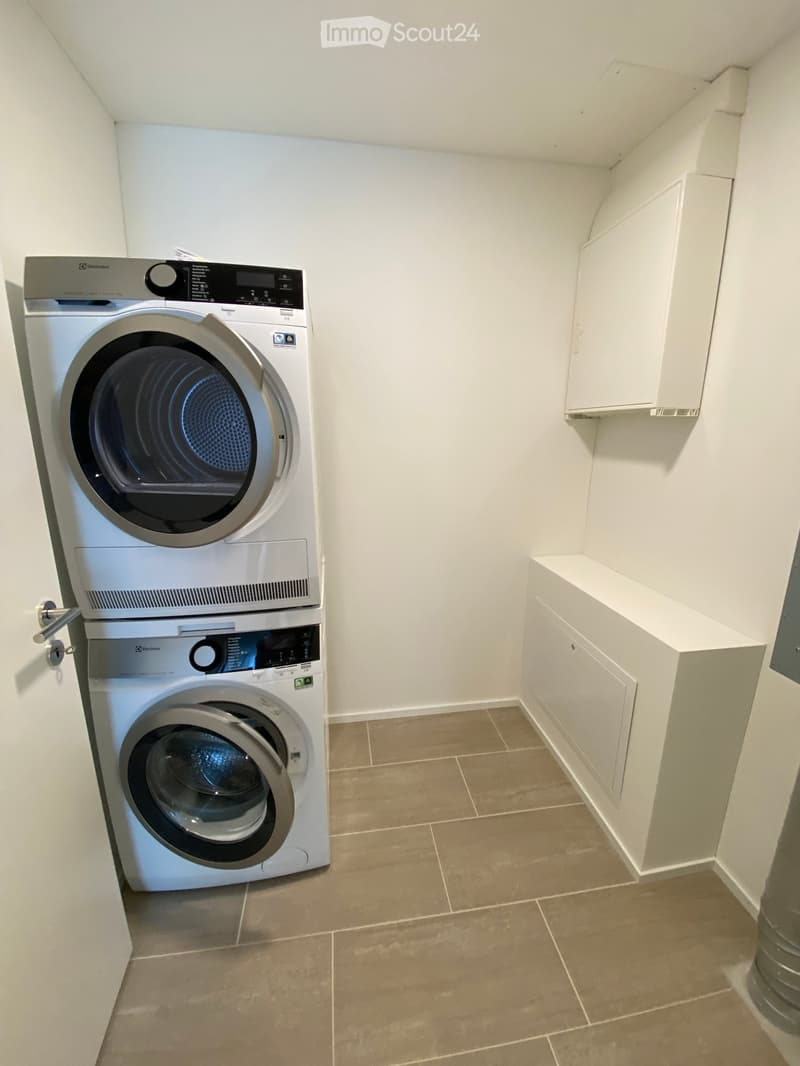A compact bathroom with a stacked washing machine and dryer unit, white tiled walls, and beige tiled flooring.