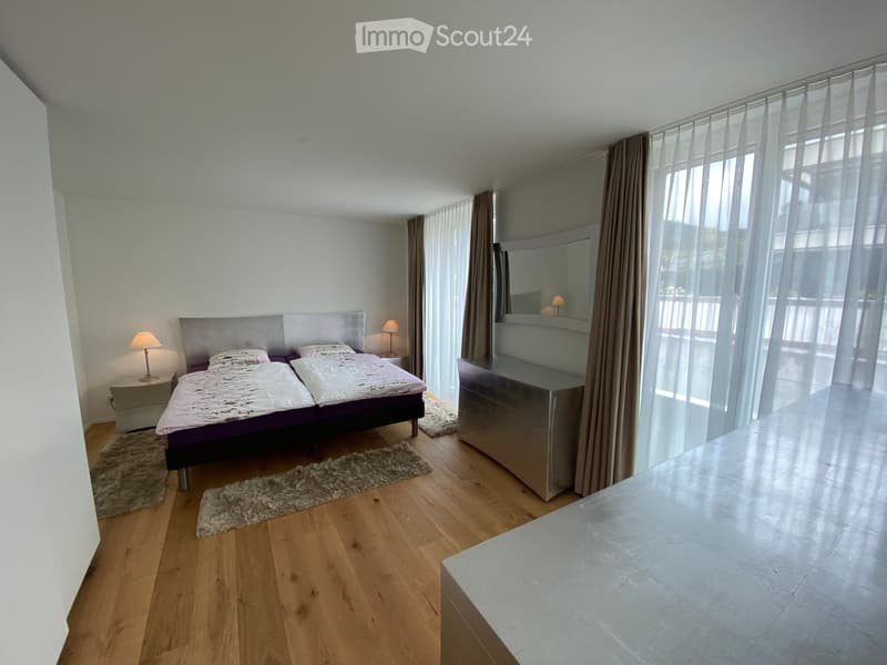 Modern bedroom with wooden flooring, large windows with sheer curtains, a double bed with a headboard, nightstands with lamps, and a mirrored vanity unit.