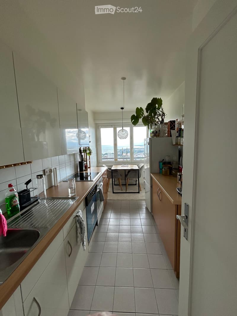 Modern kitchen with wooden cabinets, stainless steel appliances, a dining table with chairs, and a view of the city through large windows.