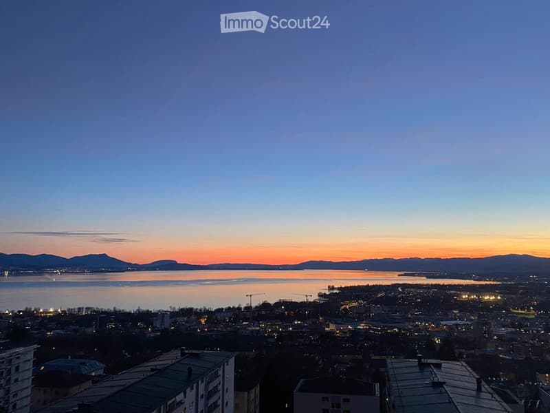 Panoramic view of a cityscape at sunset, featuring a body of water, distant mountains, and illuminated buildings.