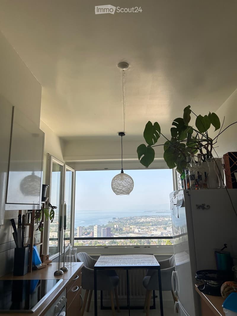 Modern kitchen with a view of the city and sea, featuring a refrigerator, dining table, chairs, pendant light, knife block, candle, and plant.