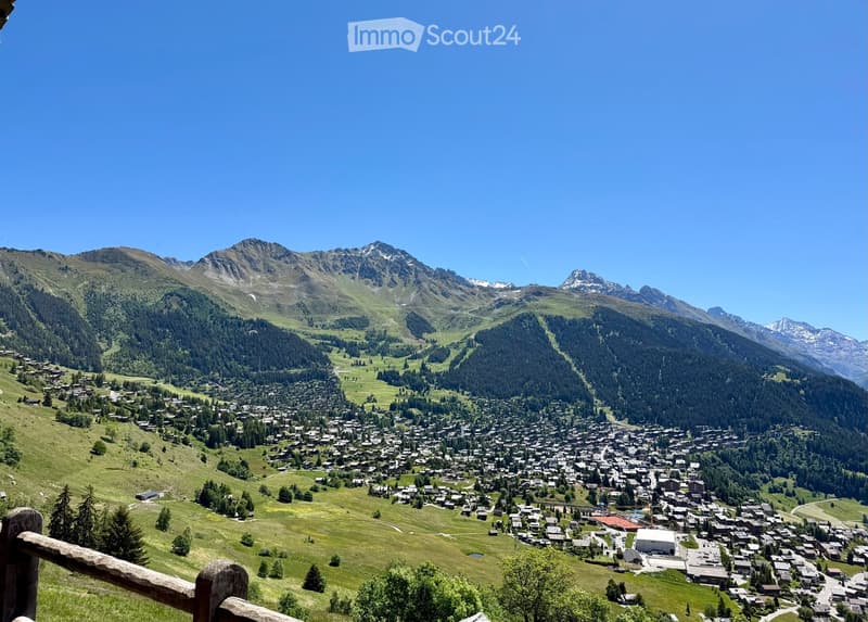 A panoramic view of a picturesque alpine village nestled in a lush green valley surrounded by towering mountains. The village features numerous houses and buildings scattered across the landscape, with a clear blue sky overhead.