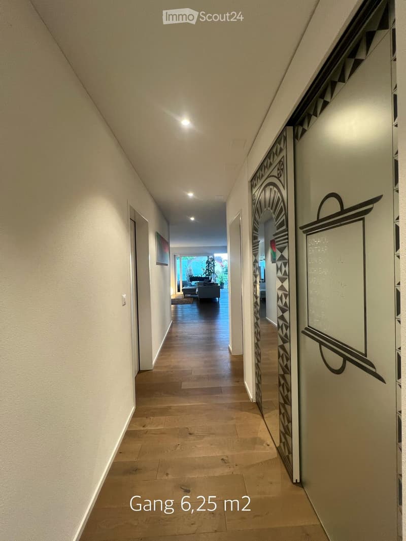 A well-lit hallway with wooden flooring, white walls, and decorative wall art. The hallway leads to a living area with a sofa and a view of a plant-filled space beyond.
