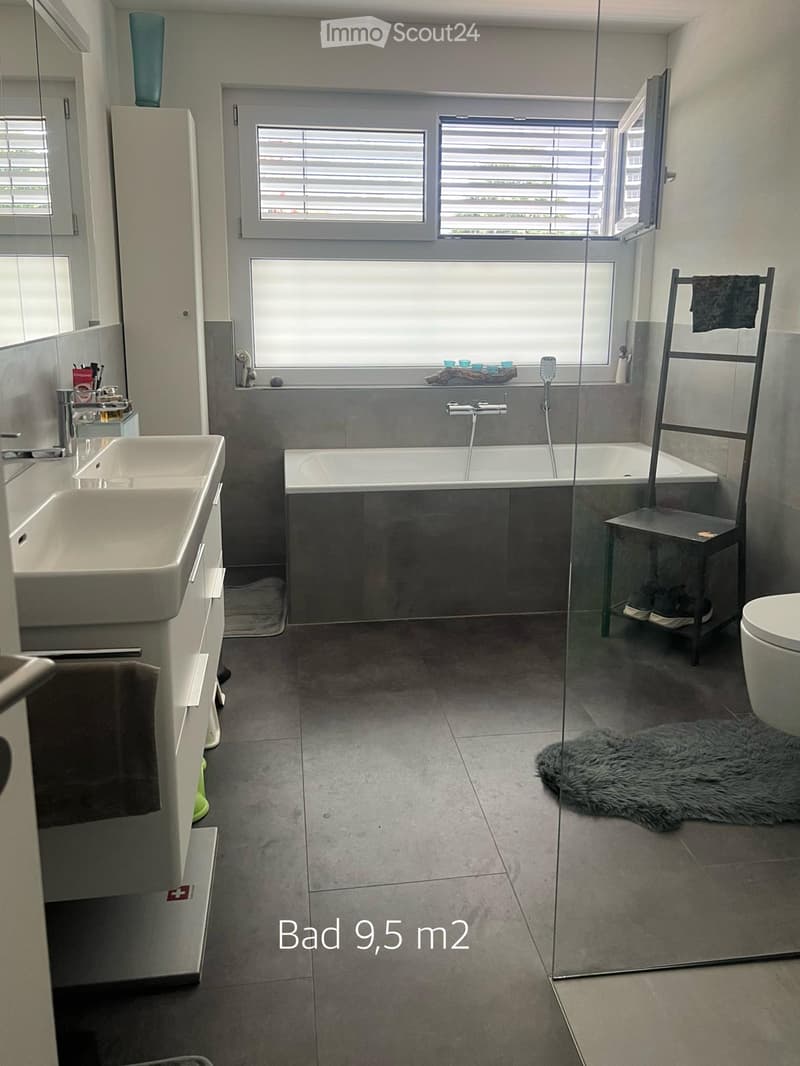 Modern bathroom with a bathtub, sink, toilet, mirror, and a towel rack. Features a large window with blinds and a fur rug on the floor.