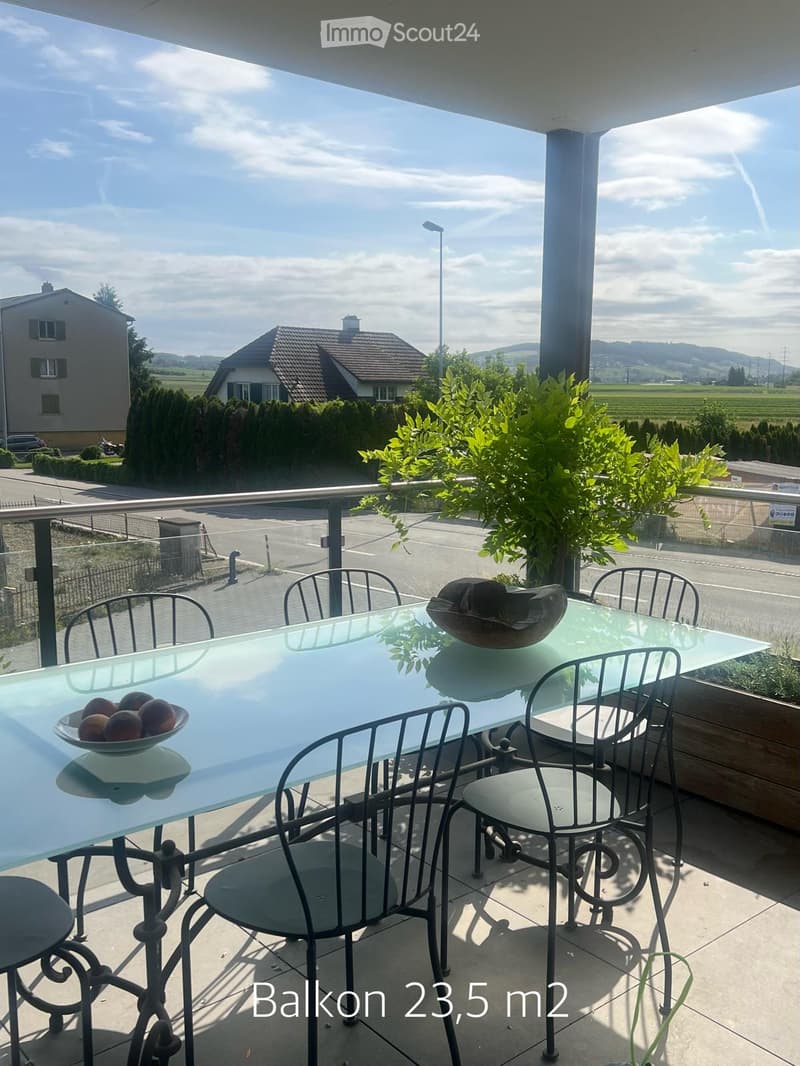Balcony with a glass table, metal chairs, and a bowl of fruit, overlooking a scenic countryside view.