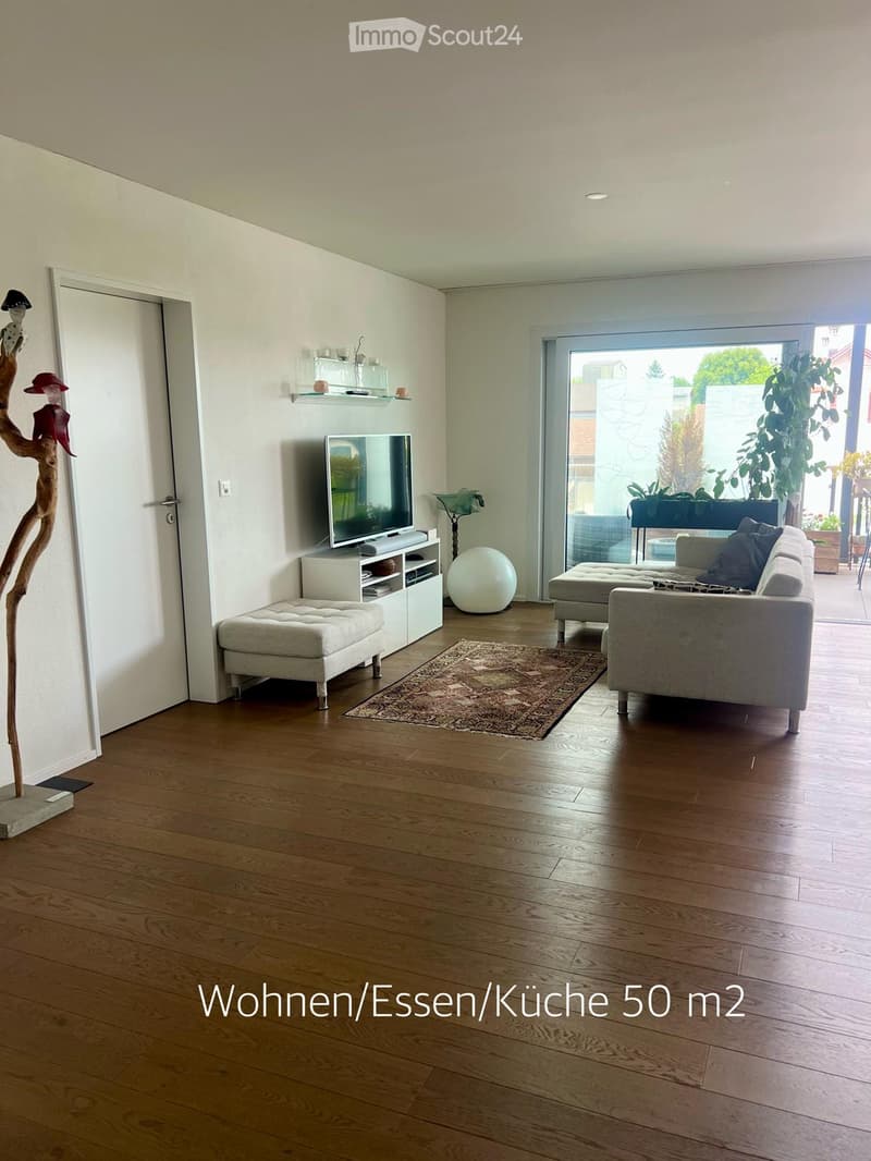 Modern living room with hardwood floors, large window, grey sofa, ottoman, coffee table, TV, decorative ball, and a unique wooden sculpture.