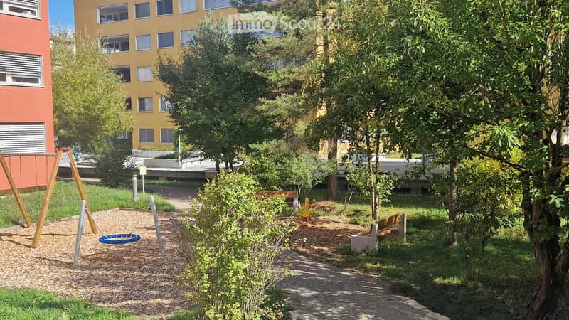 Outdoor park area with a swing set, benches, trees, and a paved pathway, surrounded by buildings.