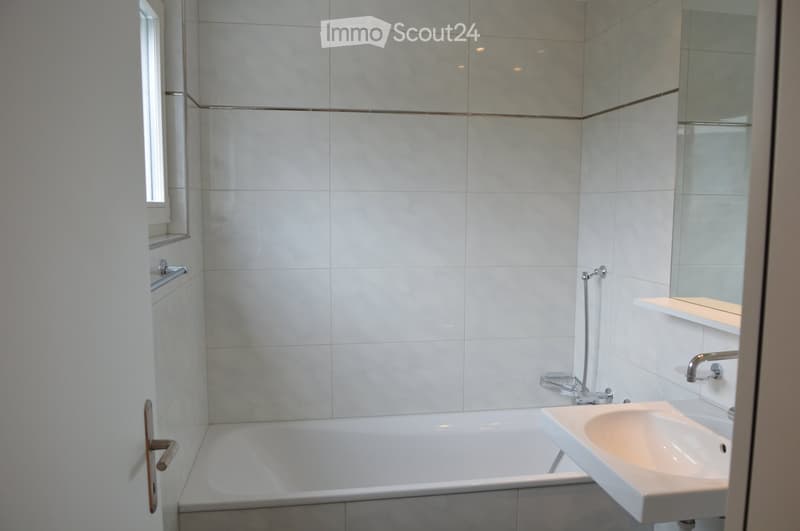 Modern bathroom with white tiles, a bathtub, a sink, a mirror, and a shelf.