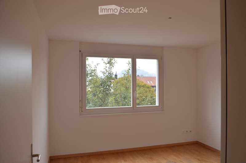 Empty room with light-colored walls, wooden flooring, and a large window offering a view of trees and a distant building.