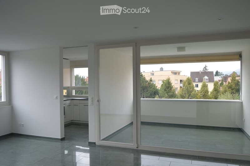 Empty room with tiled floor, white walls, and large glass sliding doors leading to a balcony with a view of trees and buildings.