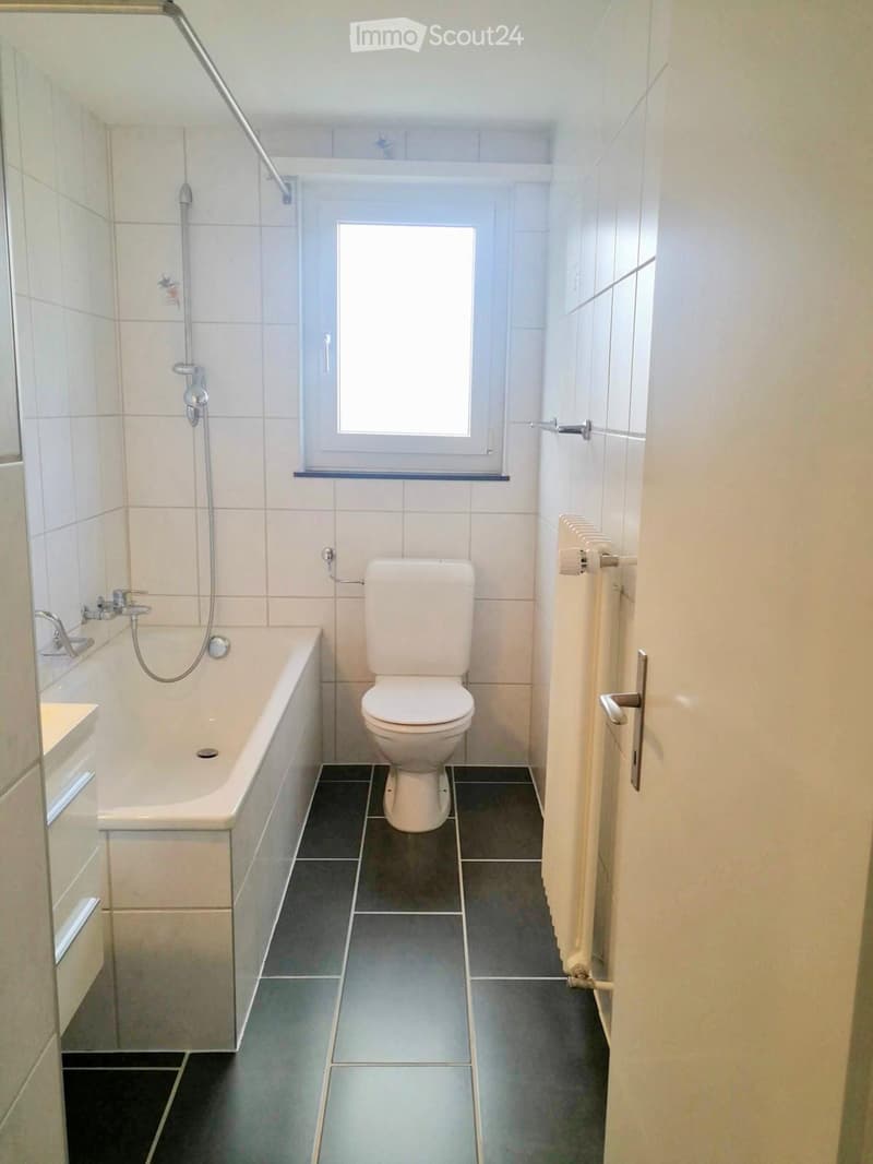 Modern bathroom with a bathtub, toilet, and showerhead. The room features white tiled walls and a dark tiled floor, with a window providing natural light.