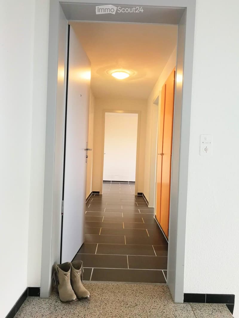 A clean, well-lit hallway with tiled floors, white walls, and recessed ceiling lighting. Two pairs of beige boots are placed near the entrance.