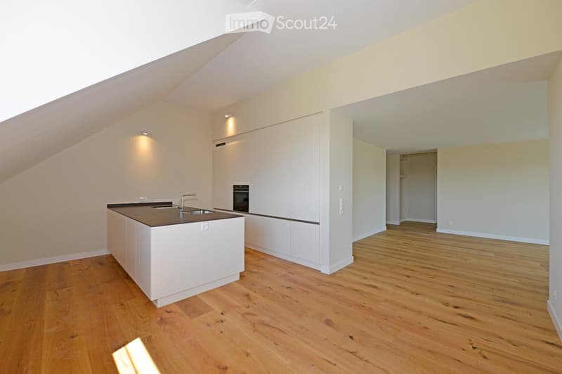 An unfurnished kitchen with white cabinets, a dark countertop, and a sink. The room features wooden flooring and a slanted ceiling with recessed lighting. An adjacent open space is visible.