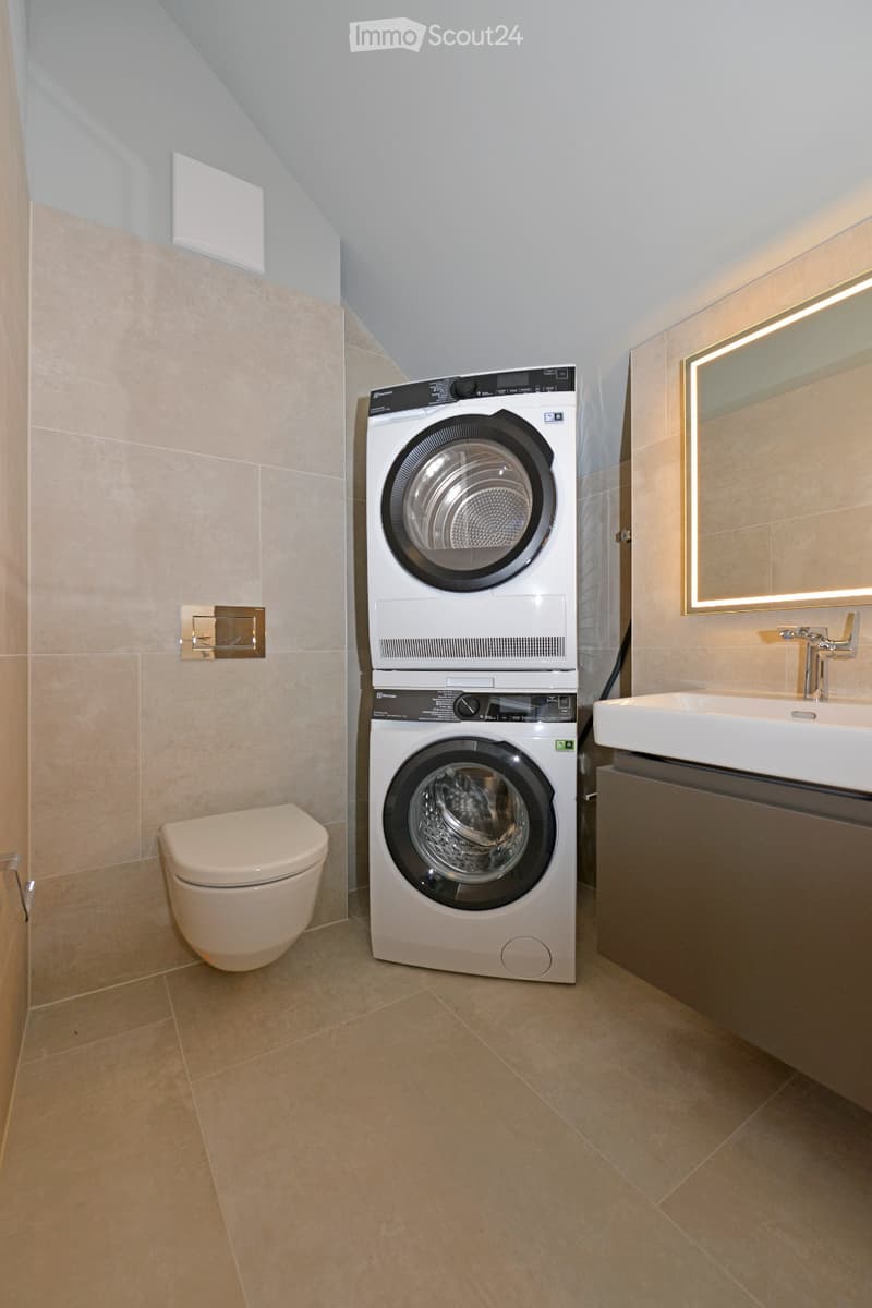 Modern bathroom with beige tiles, a toilet, a stacked washing machine and dryer, a sink with a mirror, and a sleek design.