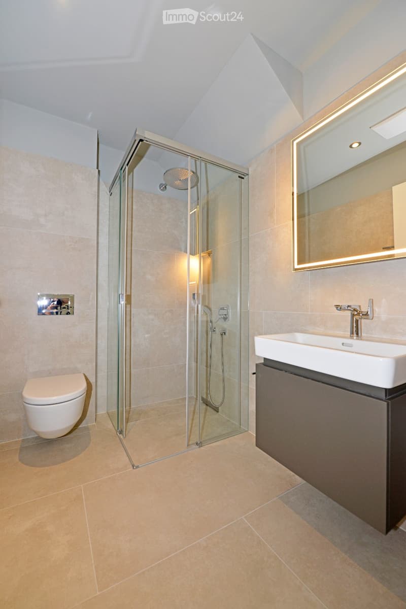 Modern bathroom with beige tiles, glass-enclosed shower, toilet, sink with modern faucet, and illuminated mirror.