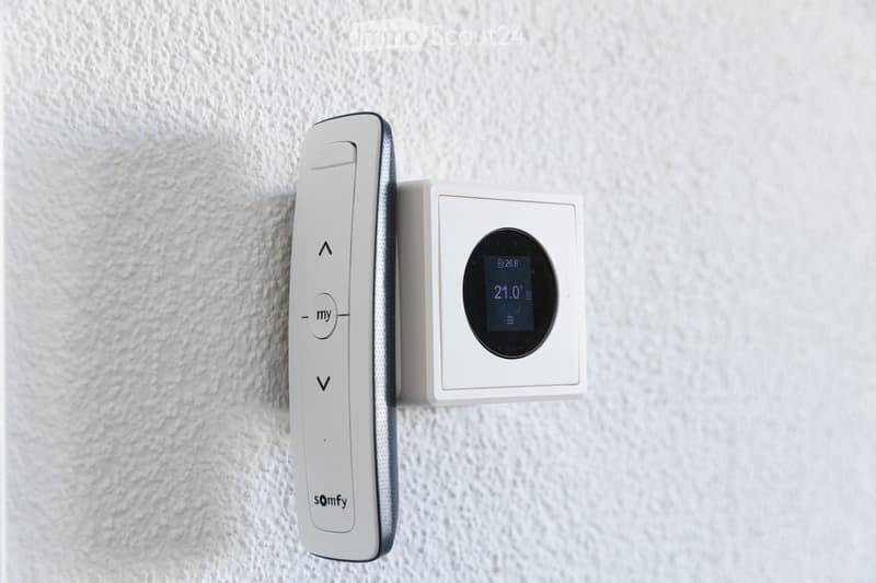 A modern white thermostat with a digital display showing temperature and a remote control attached to the wall.