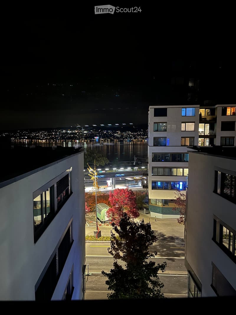 Nighttime view from a balcony showing a cityscape with illuminated buildings, a waterfront, and a train station in the distance.
