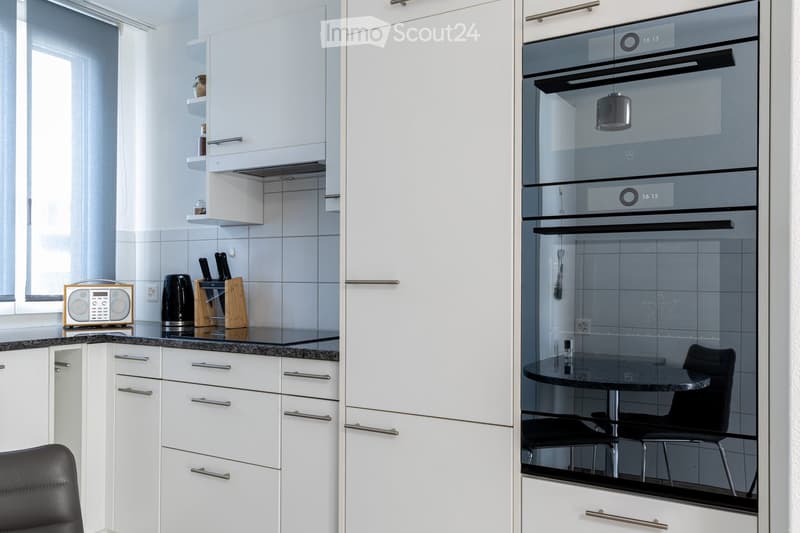 Modern kitchen with white cabinets, black countertops, and stainless steel appliances. Features include an oven, a small table with chairs, and a knife block on the counter.
