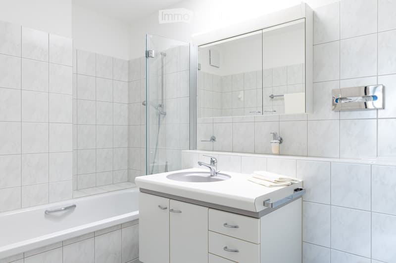 Modern bathroom with white tiles, a sink with a chrome faucet, a large mirror, a bathtub, and a glass-enclosed shower area.