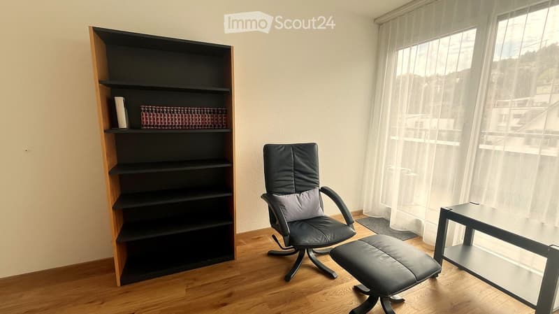 A modern, furnished room with a black leather chair, matching stool, small black table, and a tall wooden shelf with books and decorative items.