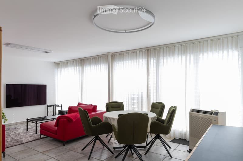 Modern living room with a red sofa, a round dining table with green chairs, a television mounted on the wall, large windows with white curtains, and an air conditioner unit.