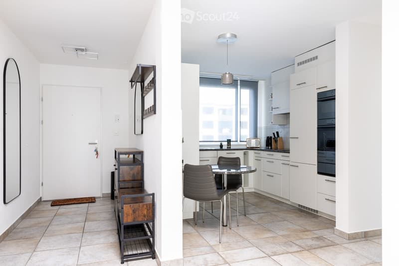 Modern, open-plan living and dining area with white walls, tiled flooring, and contemporary furniture including a dining table, chairs, and built-in cabinets.