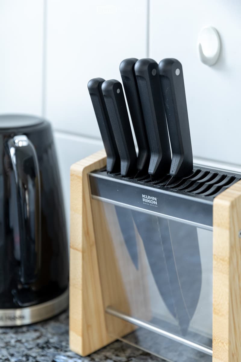 A wooden knife block with several knives inserted, placed on a marble countertop next to a black coffee maker.