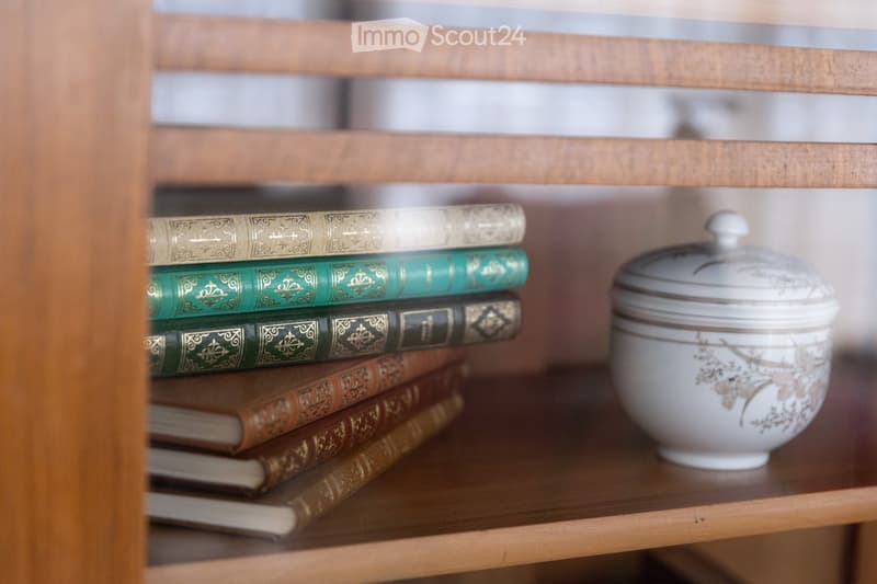 A wooden shelf displays a stack of vintage books with decorative covers and a ceramic jar with a floral pattern and lid.