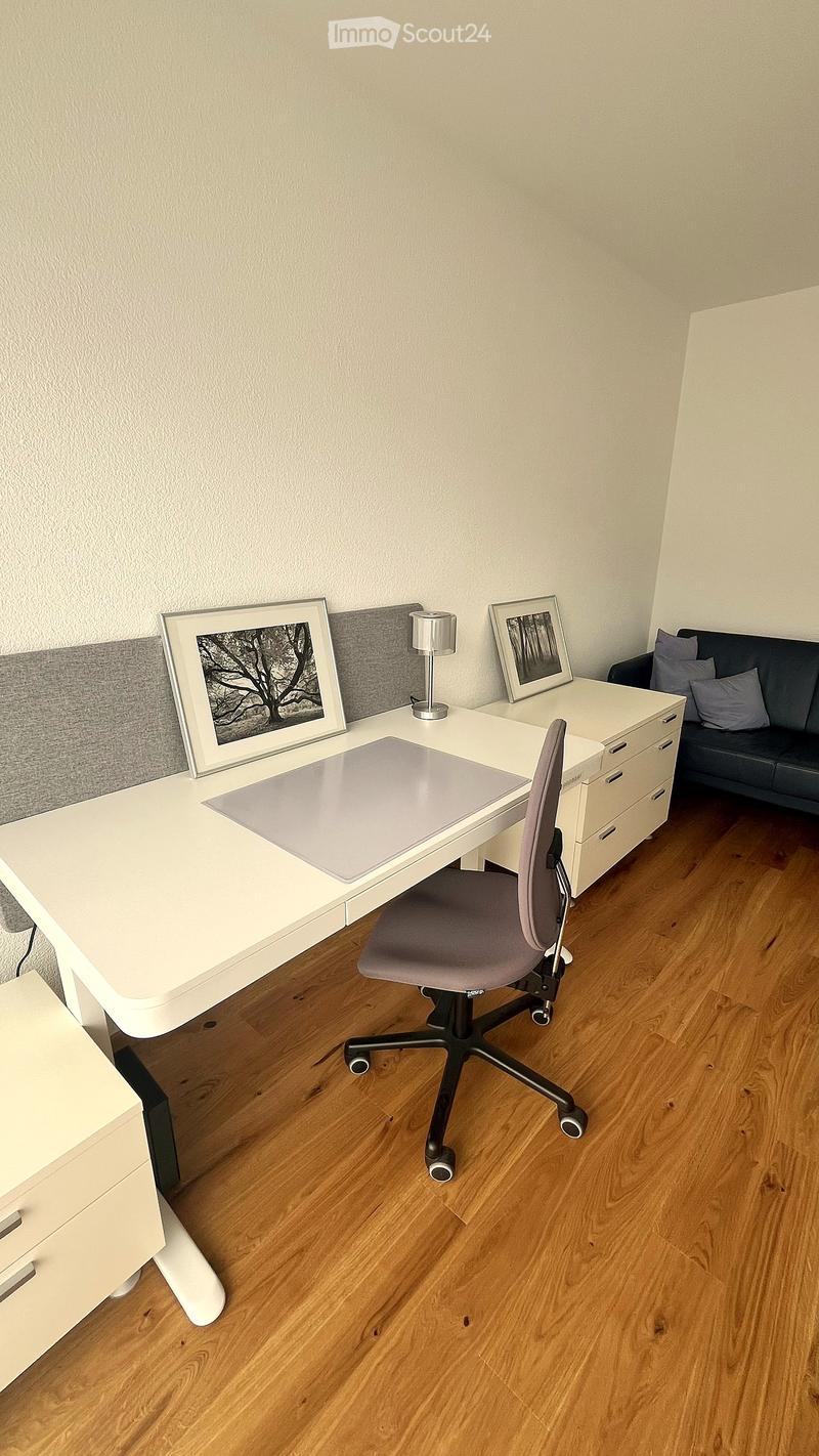 A modern, well-lit room with a white desk, ergonomic chair, grey sofa, and decorative picture frames on the wall.