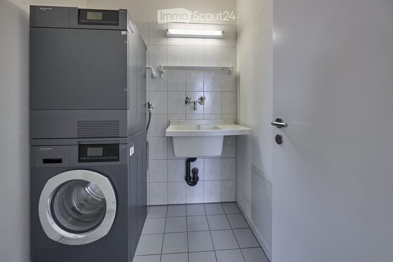 Modern bathroom with a washing machine, a white sink, and a faucet. The walls are tiled, and the floor is tiled as well.