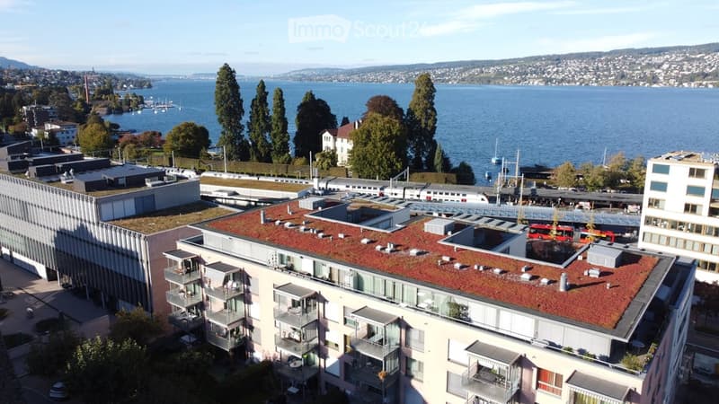 A modern building complex with a red-tiled roof and multiple balconies overlooking a scenic waterfront with a lake and distant mountains.