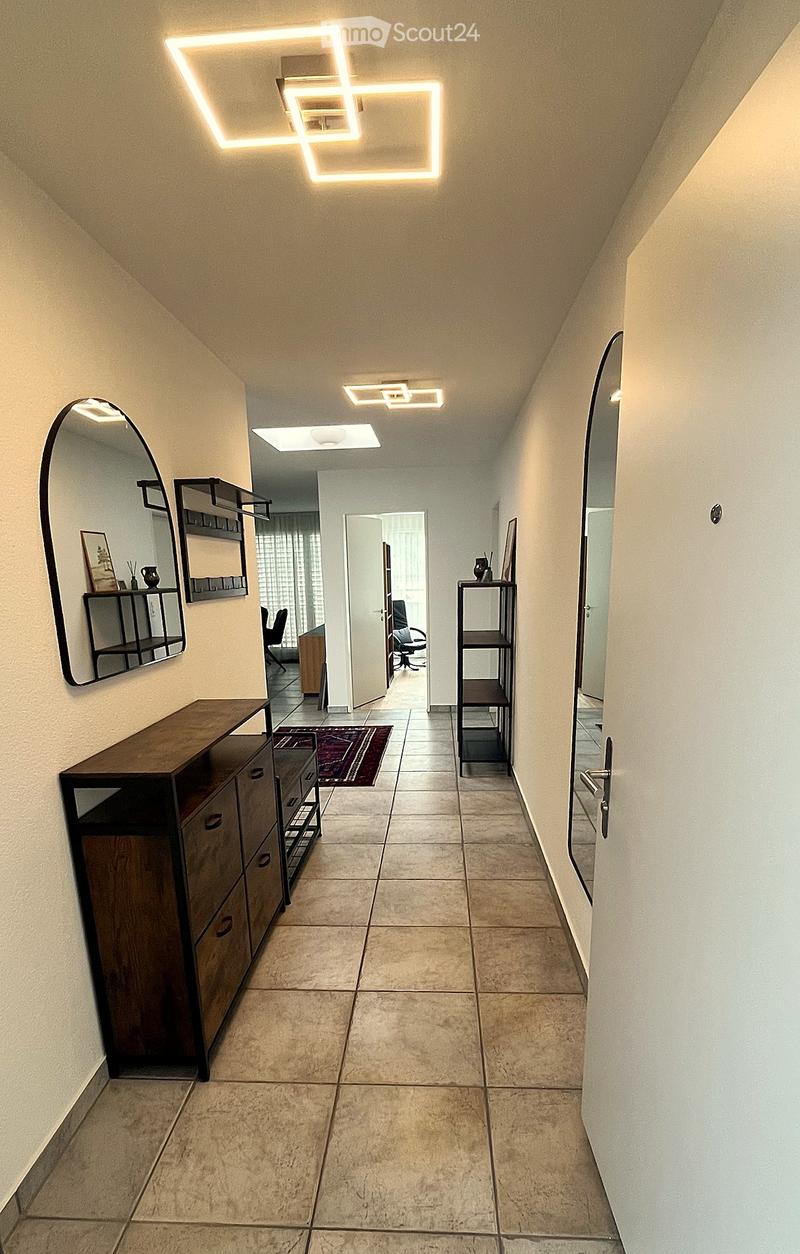 Modern hallway with beige tiled flooring, white walls, and geometric LED ceiling lights. Features a wooden cabinet, a small shelf, and a mirror on the left, with a doorway leading to another room.