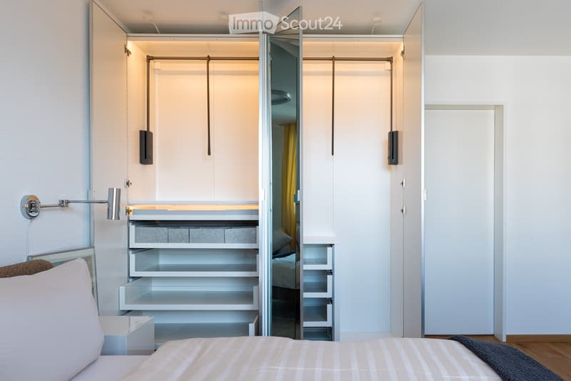 Modern bedroom with a neatly made bed, white nightstand, built-in wardrobe with mirrored doors, and a sleek wall-mounted lamp.