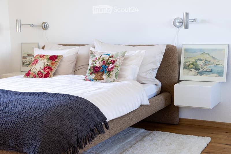 A neatly made bed with white and pink bedding, decorative pillows, and a dark blue throw blanket. A nightstand with a picture frame sits beside the bed, and wall-mounted lamps provide lighting.