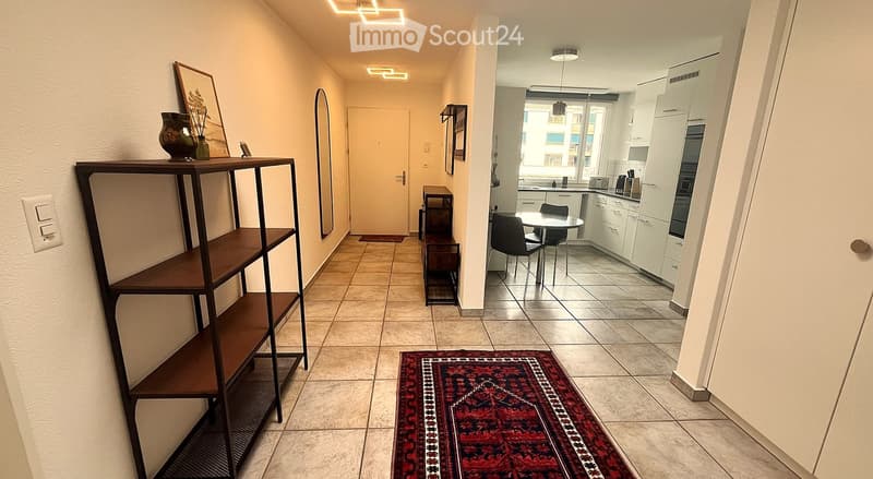 Hallway with tiled floor, wooden shelving unit, small table, and a red patterned rug leading to a kitchen area with white cabinets, a dining table, and chairs.