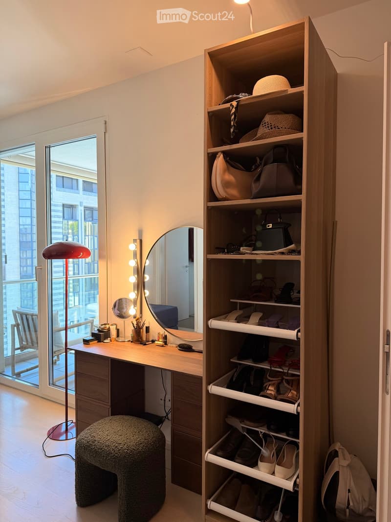 A well-organized dressing area with a vanity table, mirror, floor lamp, and a tall wardrobe filled with shoes, handbags, and hats. A round, fluffy stool is placed in front of the vanity.
